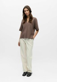 Femme debout les mains dans les poches, portant un haut marron à manches courtes, un pantalon large crème et des mocassins noirs sur un fond blanc.