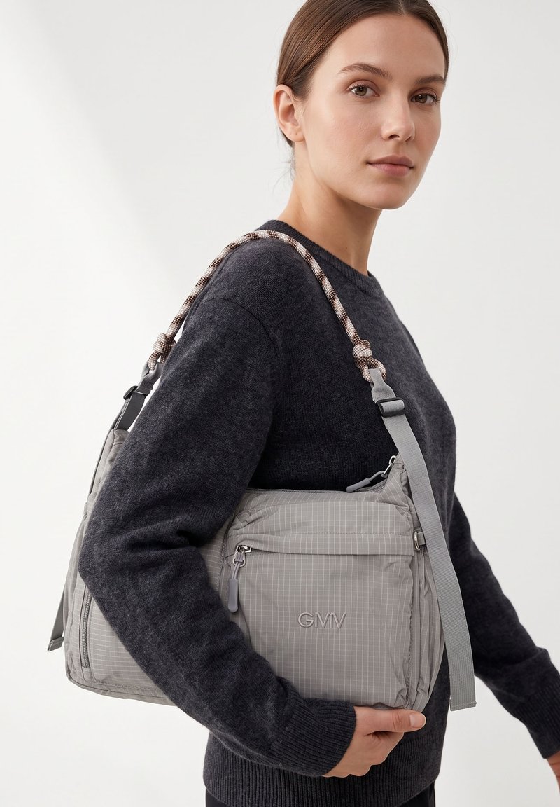 Femme portant un pull sombre porte un sac à épaule gris moyen avec poche avant zippée et bandoulière style corde sur l’épaule.