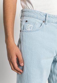 Jeans em denim azul claro com um corte relaxado, apresentando um logótipo proeminente no bolso da frente, e detalhes de ferragens metálicas visíveis e costura.