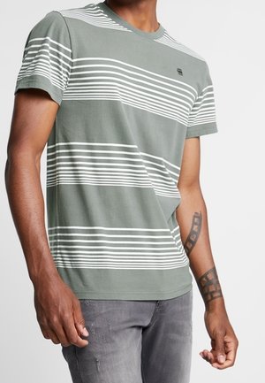 Homme portant un T-shirt vert à rayures blanches horizontales et un jean gris, montrant ses avant-bras tatoués sur un fond uni.