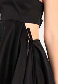 Schwarzes Kleid mit einem taillierten Oberteil, schwingendem Rock, seitlichem Schlitz und einer Bindung in der Taille. Hergestellt aus glattem, leichtem Stoff.