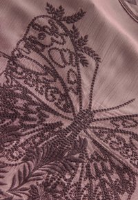 Un tissu brodé dans un mauve doux présente un design de papillon détaillé avec des motifs et des textures complexes, renforçant la profondeur et la dimension visuelles.