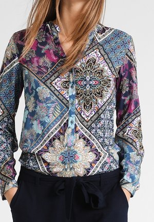 Vrouw draagt een blouse met lange mouwen en knopen met meerkleurige, ingewikkelde bloem- en geometrische patronen, ingestopt in een zwarte broek.