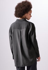 Camicia leggera di un grigio scuro, con una texture stropicciata, maniche lunghe e polsini con bottone. Presenta un colletto classico e un orlo leggermente curvo.