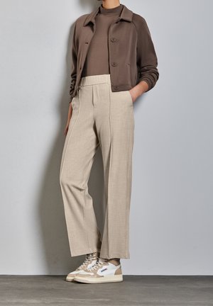 Ruskea lyhyt takki ruskean topin päällä, yhdistetty vaaleanbeigeihin räätälöityihin housuihin sekä beigen ja valkoisen sävyisiin lenkkareihin. Sileät tekstuurit ja puhtaat linjat.