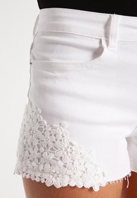 Bílé džíny šortky s detailním bílým krajkovým lemem po dolním okraji. Klasický pětizásobní design s zipem a knoflíkovým zapínáním.