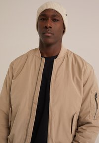 Béžová bombarďácká bunda se zipem vpředu a bočními kapsami, v kombinaci s černým tričkem s kulatým výstřihem a světlým pleteným čepicím.