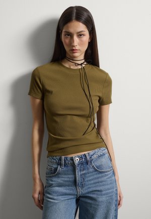 Frau mit langem, dunklem Haar, die ein eng anliegendes olivgrünes Kurzarmshirt, blaue Jeans und ein dünnes schwarzes Halsband trägt, das zu einer Schleife gebunden ist.