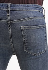 Pantalones vaqueros de mezclilla azul oscuro con una textura suave, con dos bolsillos traseros y costuras estándar a lo largo de las uniones. Cinturilla con trabillas para cinturón.