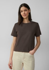 T-shirt en coton marron à manches courtes et col rond, avec une coupe décontractée et une texture lisse. Porté avec un pantalon de couleur claire.