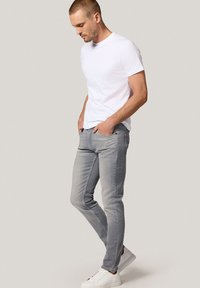 Mężczyzna w białej koszulce, szarych dopasowanych dżinsach i białych trampkach, stojący z rękami w kieszeniach, patrzący w dół na jednolitym tle.