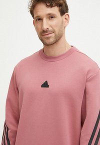 Felpa rosa con collo rotondo e logo nero Adidas sul petto. Presenta strisce nere lungo le maniche e un tessuto morbido e strutturato.