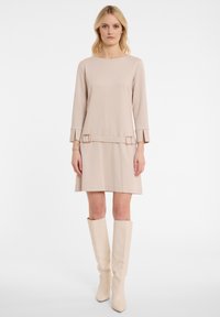 Frau in einem beigen Langarmkleid mit Schnallen-Details an der Taille und passenden kniehohen Stiefeln, die vor einem weißen Hintergrund steht.