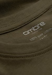 Olivegrüner Baumwoll-T-Shirt-Stoff mit glatter Textur und sichtbaren Nähten sowie einem Etikett mit der Aufschrift "100 % reine Baumwolle" und dem Markennamen.