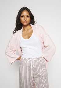 Light pink, textured kimono-style cardigan with wide sleeves, worn over a white tank top and striped, loose-fitting pants.