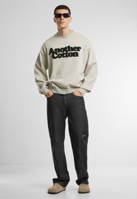 Világos bézs pulóver fekete "Another Cotton" felirattal, sötét farmernadrággal és bézs slip-on cipőkkel párosítva. A design laza és kényelmes.