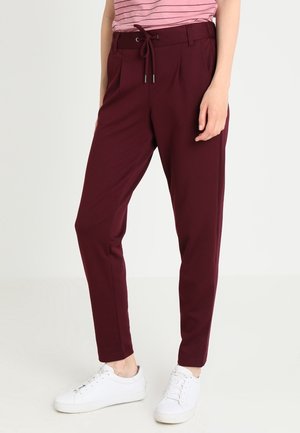 Pantalons bordeaux ajustés avec taille élastique, plis avant et tissu doux ; associés à des baskets blanches pour un look décontracté.