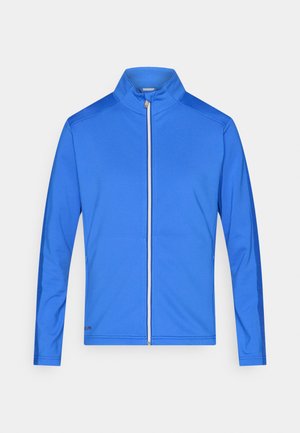 Chaqueta azul con cremallera y cuello alto, fabricada con tela ligera, que presenta una textura suave y paneles laterales más oscuros en contraste.