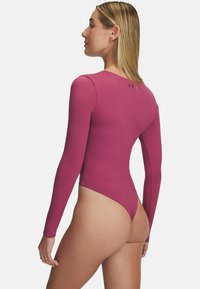 Model wearing a long-sleeve, fitted magenta swimsuit, facing away, with a minimalistic background emphasizing the clothing design.