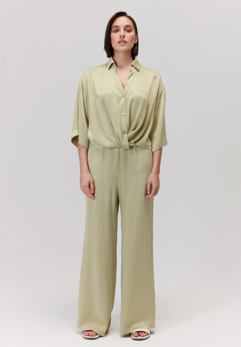 Jasnozielona, oversize koszula zapinana na guziki, związana w pasie, w połączeniu z szerokimi spodniami w tym samym kolorze, wykonanymi z gładkiego materiału.