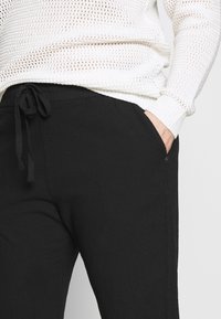 Czarne spodnie z miękkiego, teksturowanego materiału, z regulowanym paskiem ze sznurkiem w talii oraz bocznymi kieszeniami. Zbliżenie na detale tkaniny obok białego dzianinowego swetra.