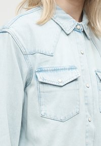 Φανελένιο πουκάμισο τζην ανοικτού μπλε με κουμπιά τύπου snap και δύο τσέπες στο στήθος, φορεμένο από άτομο με ξανθά μαλλιά.