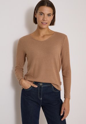 Femme aux cheveux courts et bruns portant un pull beige côtelé à manches longues et un jean bleu foncé, debout avec une main dans la poche.