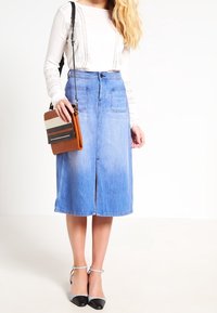 Jupe en denim avec une fente sur le devant, dotée de deux poches, d'un délavage clair et d'une coupe décontractée. Associée à un haut crème et un sac à main multicolore.