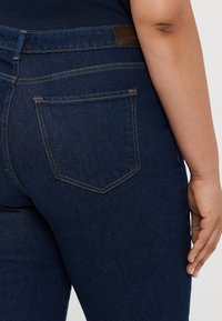 Temno modri denim kavbojke imajo srednje visok pas, zadnje žepe in kontrastne šive. Tkanina ima gladko teksturo in minimalne poškodbe.