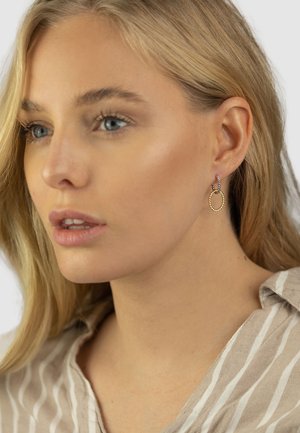 Goldene Creolen mit gedrehter Textur und einem kleinen lila Akzent. Getragen von einer Person mit hellblondem Haar und einem Oberteil in neutralen Farben.