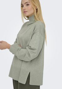 Suéter de cuello alto verde claro con puños y dobladillo acanalados, con aberturas laterales. Textura suave y corte relajado, adecuado para superponer.