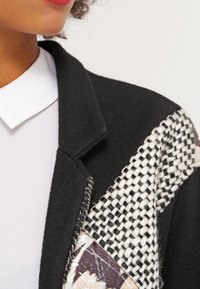 Veste en laine noire avec un motif tissé texturé blanc et noir sur une épaule, rehaussée d'un détail en tissu à motif contrasté.