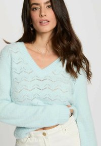 Maglione azzurro chiaro a scollo a V, lavorato a maglia, con motivo ondulato e texture trasparente. Maniche lunghe, polsini aderenti, indossato con pantaloni bianchi a vita alta.
