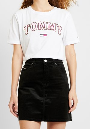 Kvinde iført en hvid t-shirt med teksten og logoet "TOMMY", kombineret med et sort denimskørt med høj talje, stående foran en ensfarvet baggrund.