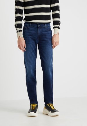 Mann trägt dunkelblaue, eng anliegende Jeans, schwarzen Pullover mit weißen horizontalen Streifen und schwarze klobige Sneaker mit gelben Schnürsenkeln.