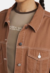 Giacca di jeans marrone con accessori in argento, due tasche sul petto e cuciture bianche a contrasto, indossata sopra una maglietta beige a coste.