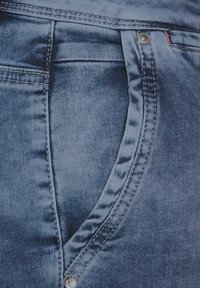 Primer plano de una tela de mezclilla que presenta un color azul oscuro, costuras visibles, detalles de bolsillo y un acento de botón plateado. Se observa una textura suave.
