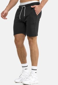 Mann in schwarzen Shorts mit Kordelzug, weißen Sportsocken mit schwarzen Streifen und weißen Turnschuhen, der mit einer Hand in der Tasche steht.