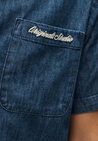 Τσέπη από τζιν μπλε πουκάμισο με λευκό κεντητό κείμενο «Originals Studio» στο πάνω μέρος της ραφής της τσέπης.