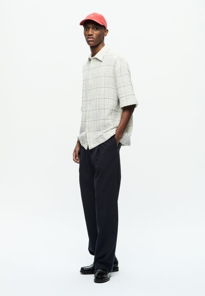 Chemise à manches courtes en carreaux gris clair avec des boutons dorés, associée à un pantalon bleu foncé et des mocassins noirs. Une casquette rouge complète la tenue.