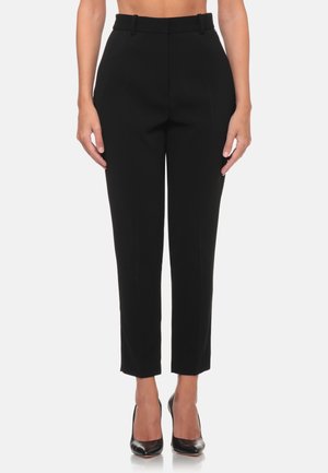 Donna che indossa pantaloni neri aderenti a vita alta alla caviglia e scarpe nere con tacchi alti, in piedi su uno sfondo bianco uniforme.