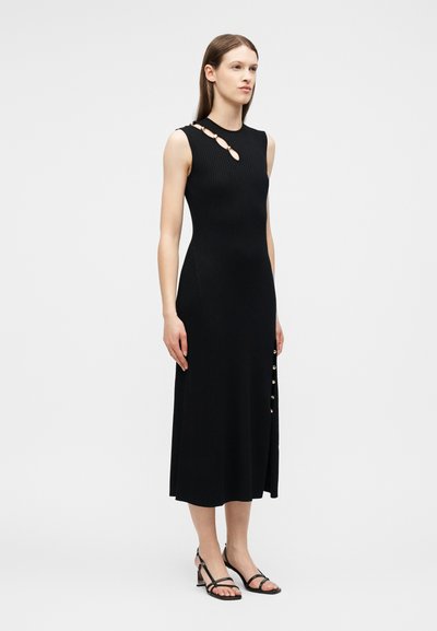 Femme portant une robe midi noire sans manches avec des détails de bagues dorées sur la poitrine et les côtés, associée à des sandales noires à talons blocs et brides.