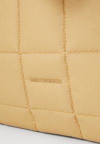 Gros plan sur un tissu matelassé beige avec des coutures visibles et le nom de la marque ARKK Copenhagen embossé au centre.