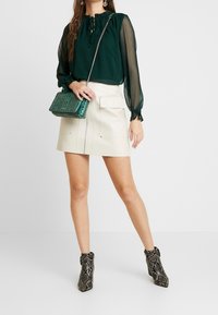 Camicia verde trasparente con maniche lunghe a sbuffo, minigonna in ecopelle crema con zip anteriore e stivaletti alla caviglia in stampa serpente. Borsa verde con texture.