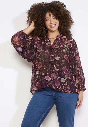 Blusa transparente con patrón floral, mangas largas, fondo de color púrpura intenso, cierre de botones y corte holgado, combinada con vaqueros azules.