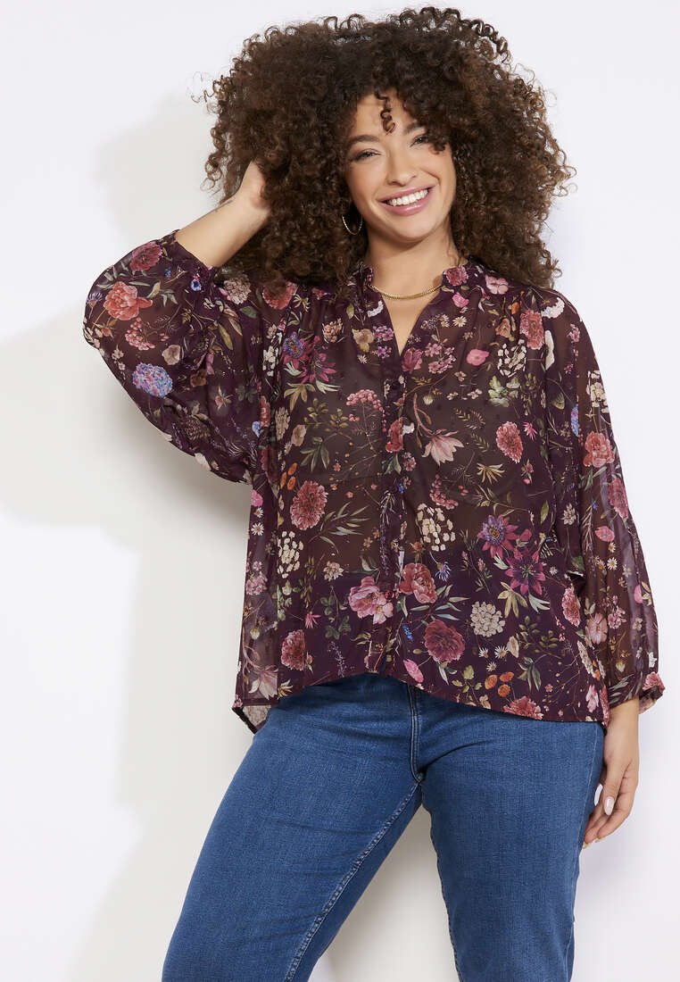 Blusa trasparente con motivo floreale, maniche lunghe, sfondo viola profondo, chiusura con bottoni e vestibilità comoda, abbinata a un paio di jeans blu.