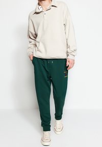 Beige svetr s límcem a knoflíky, doplněný tmavě zelenými tepláky s vyšitým vzorem. Světle barevné tenisky outfit doplňují.