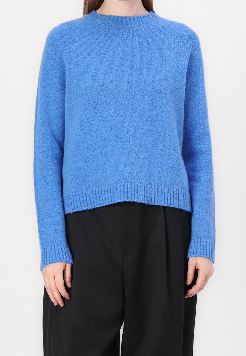 Personne portant un pull bleu doux à manches longues associé à un pantalon noir ample, debout sur un fond clair uni.