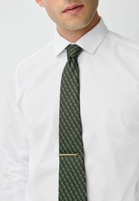 Green patterned tie made of textured fabric, displaying a diagonal design with white accents, secured with a gold tie bar.