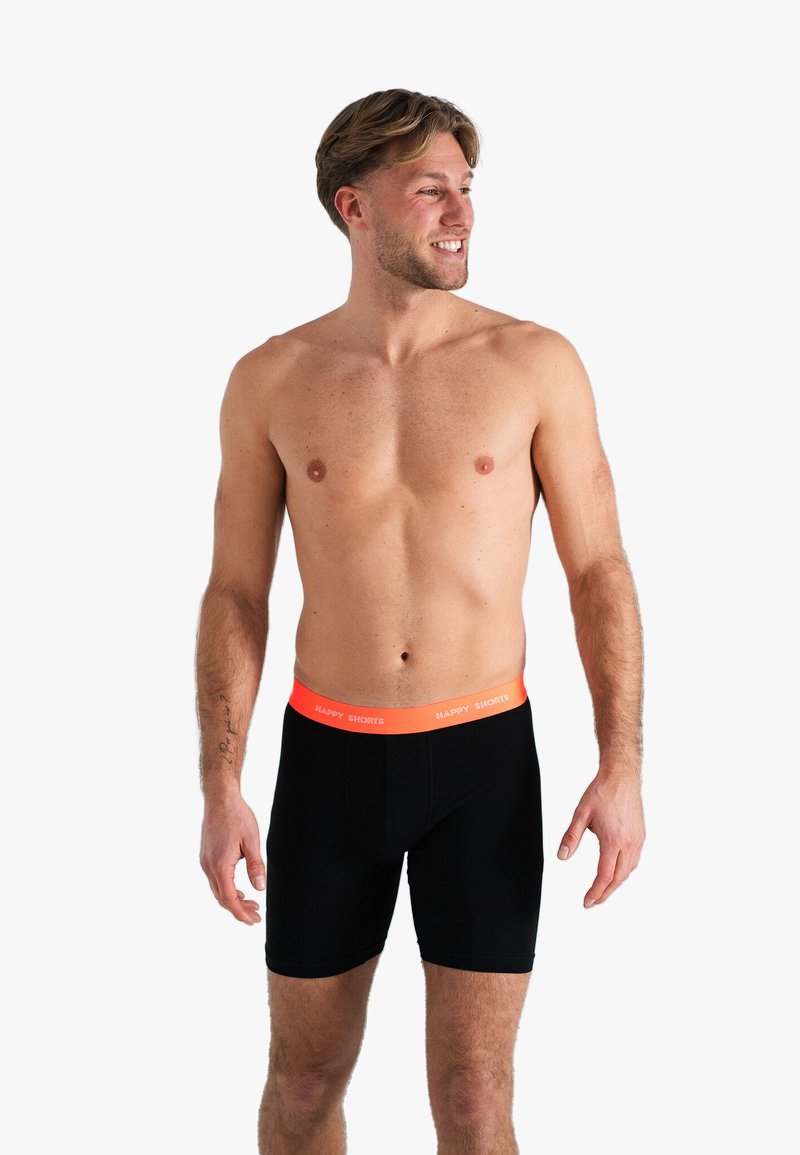 Zwarte boxershorts met een elastische oranje tailleband met het label "HAPPY SHORTS." Het materiaal lijkt zacht en rekbaar, ontworpen voor comfort.